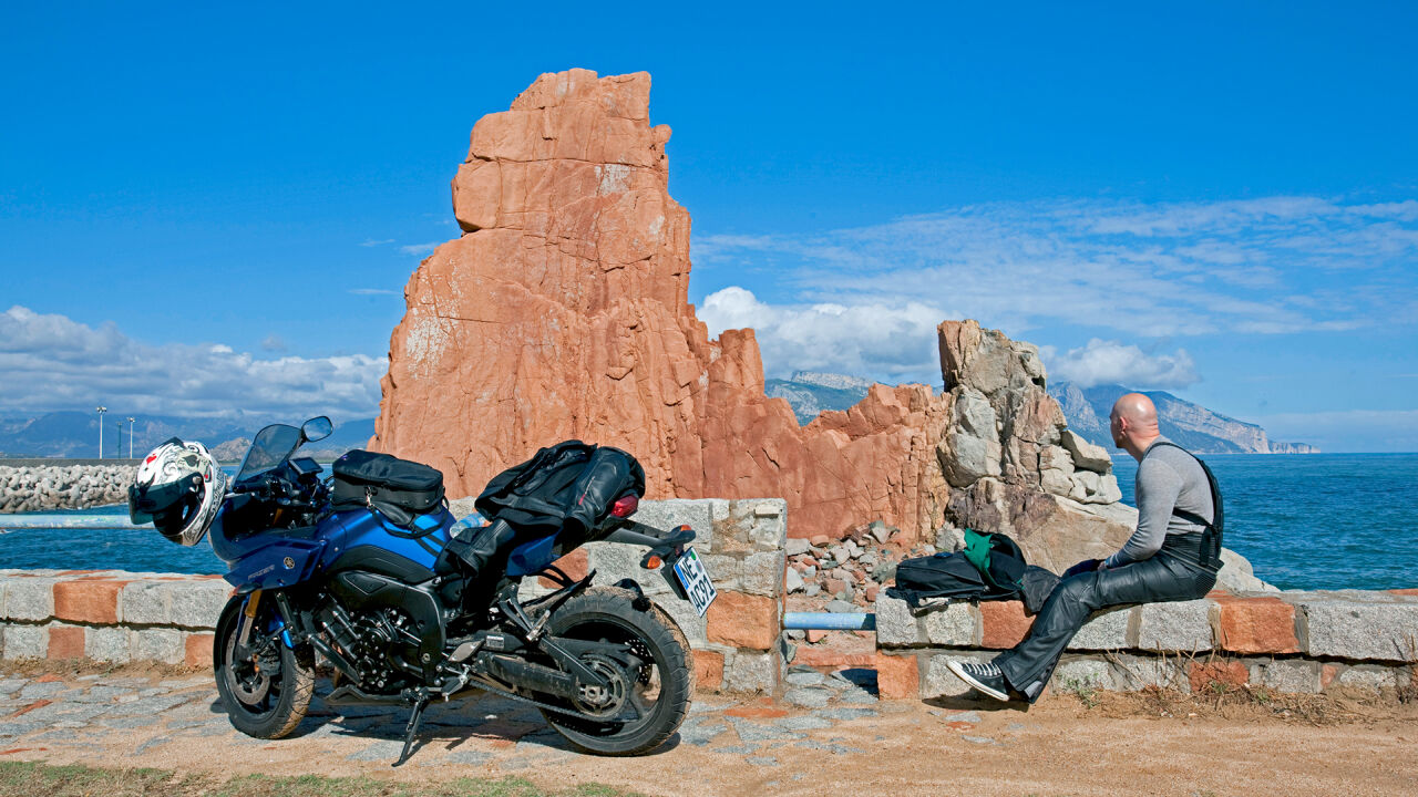 Roter Porphyrfelsen am Meer, davor ein geparktes Motorrad und ein Fahrer auf einer Mauer